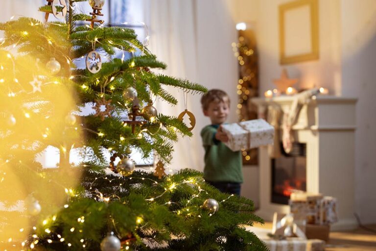 Ein kleiner Junge trägt ein Geschenk zum geschmückten Weihnachtsbaum.