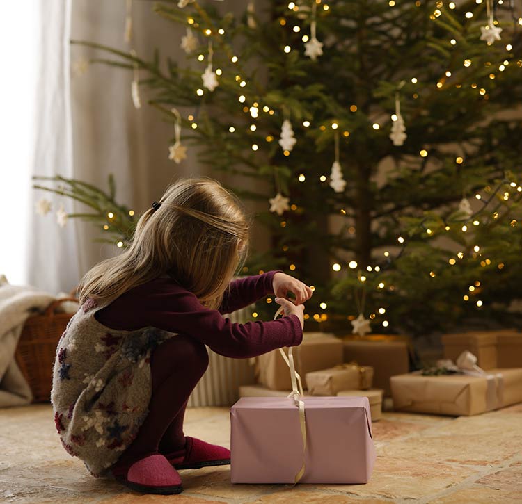 Ein kleines Mädchen sitzt vor dem Weihnachtsbaum und packt ein Geschenk aus.