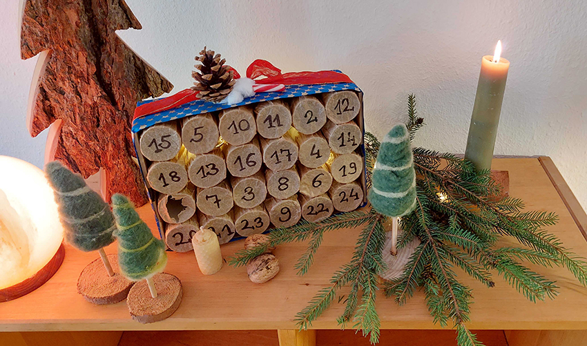 Der Adventskalender aus Klopapierrollen steht auf einer Holzkommode und ist weihnachtlich dekoriert.