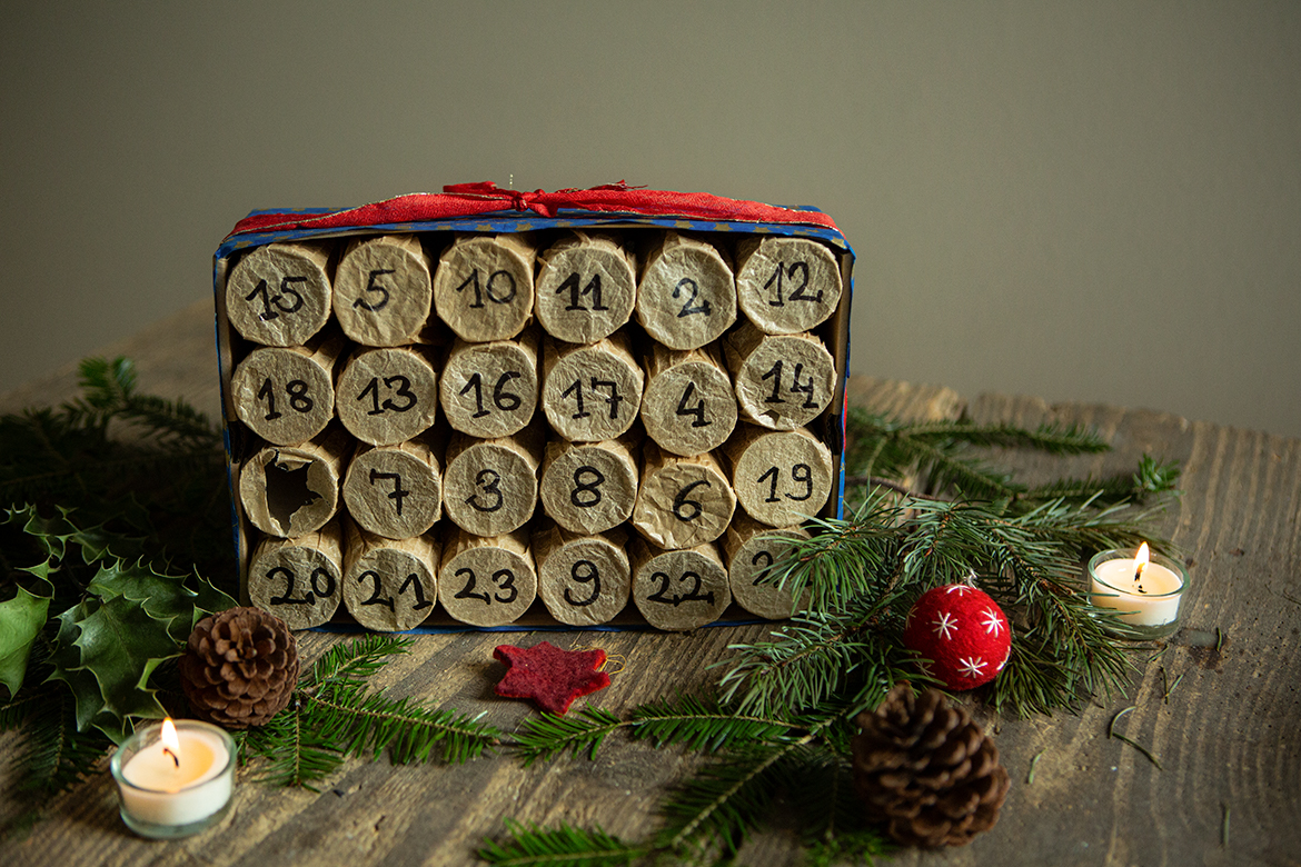 Der Adventskalender aus Klopapierrollen steht auf einem Holzbrett, um den Kalender herum liegen Tannenzweige und Weihnachtsschmuck.