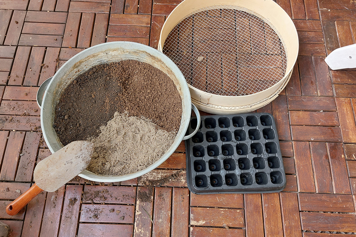 Zum Anzuchterde selbst machen steht ein Eimer mit Sand, Kompost und Erde auf dem Boden, daneben ein Sieb und eine Schaufel.