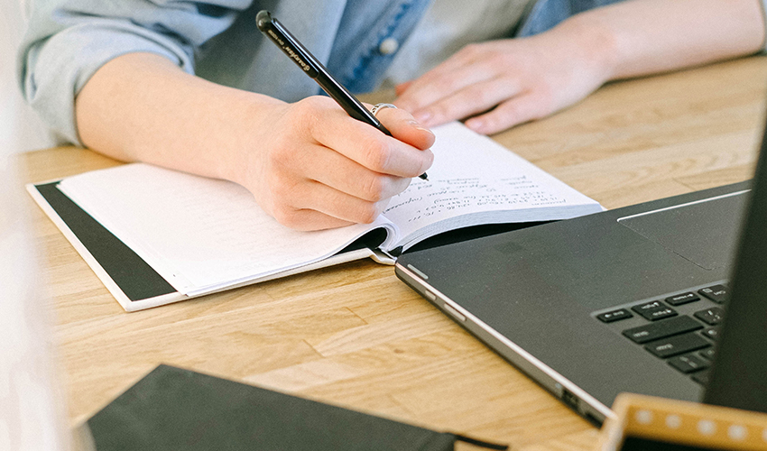 Frau sitzt mit einem Notizbuch und einem Stift in der Hand vor ihrem Laptop und schreibt eine Aufgabenliste ins Buch.