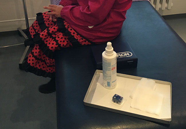 Das Mädchen sitzt auf einer Liege, neben sich steht ein tablett mit den Piercingutensilien.