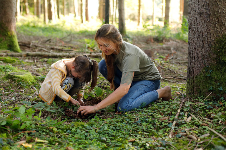 Eine Frau kniet mit ihrer Tochter auf dem Waldboden und ist mit Blätter bestimmen beschäftigt.