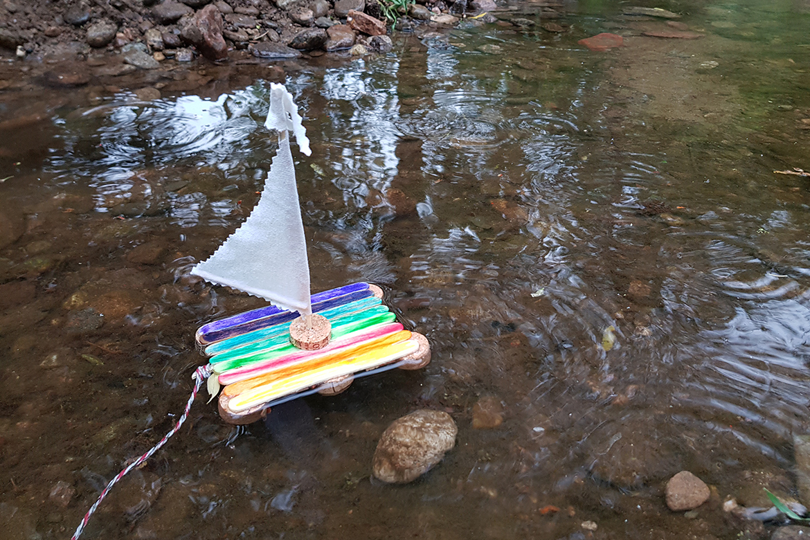 Ein gebasteltes Spielzeug aus Eisstielen, hier ein Boot, das bunt angemalt auf einem Bach fährt.