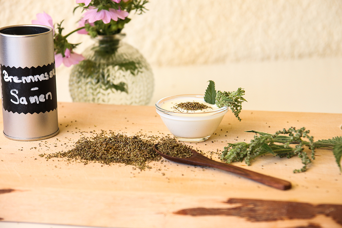 Gesammelte Brennnesselsamen liegen auf einem Holztablett, auf dem auch ein Schälchen mit Joghurt und Brennnesselsamen steht.