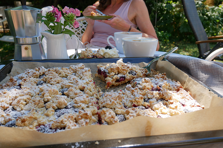 Das Blech mit dem Brombeerkuchen steht auf einem gedeckten Kaffeetisch, im Hintergrund isst eine Frau eine Stück des Kuchens.