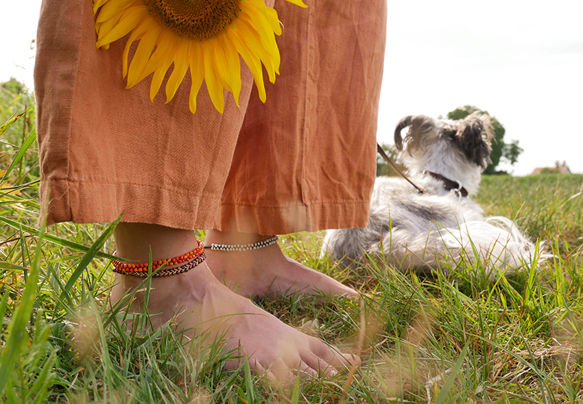 Die Frau steht mit ihrem Hund auf einer Wiese und trägt an den Fußgelenken gleich mehrere bunte Fußkettchen.