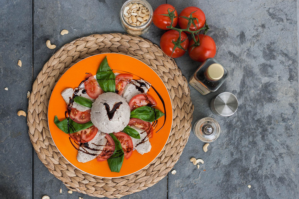 Lecker angerichtet liegt der vegane Mpzarella in Scheiben geschnitten zusammen mit Tomaten und Basilikum auf dem Teller.