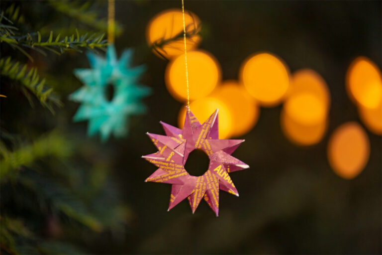 Im Vordergrund hängt ein pinker, im Hintergrund ein türkiser Deko-Stern an einem Tannenbaum