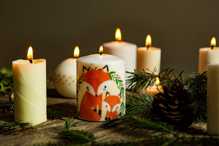 Viele brennende Kerzen stehen auf einem Tisch, die vorderste Kerze ist mit einem Fuchsmotiv verziert.