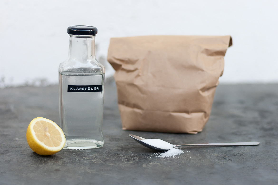 Eine aufgeschnittene Zitrone, eine Flasche Klarspüler und eine braune Papiertüte stehen auf grauer Fläche.