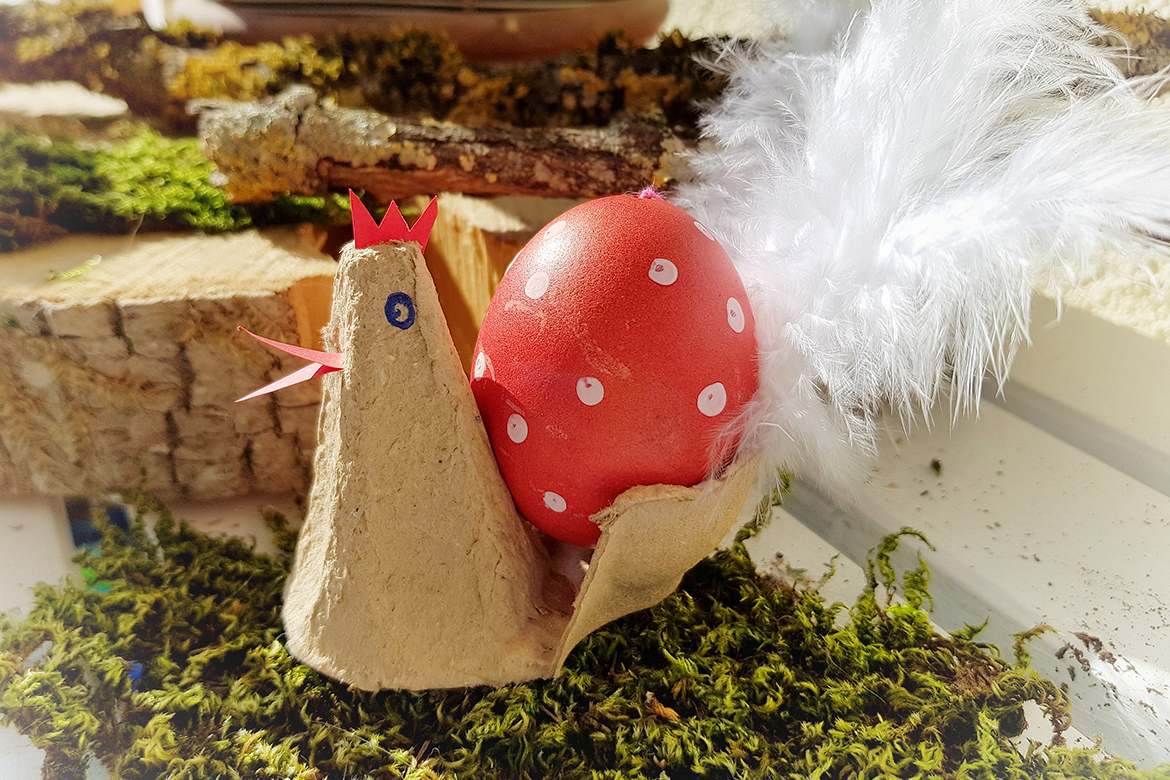 Das Hähnchen sitzt auf Moos und trägt in rotes Ei mit weißen Punkten.