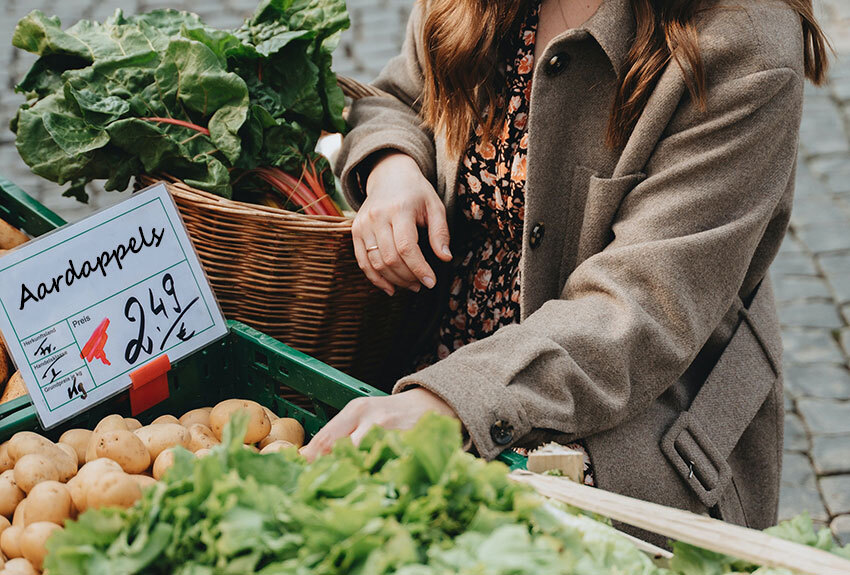 Een vrouw met een boodschappenmand aan haar arm koopt groenten op de markt volgens de seizoenskalender.