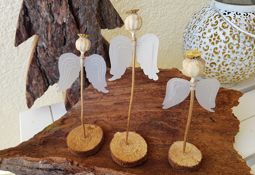 Drie engelen gemaakt van papaverbollen staan op een houten schijf.