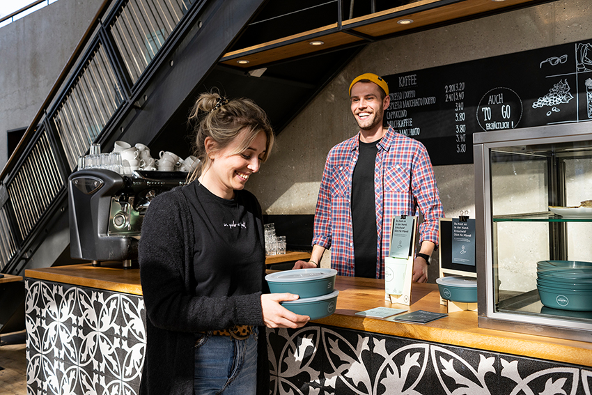 An einer Restauranttheke nimmt eine Frau Essen-to-go in Mehrwegschalgen von RECUP mit.