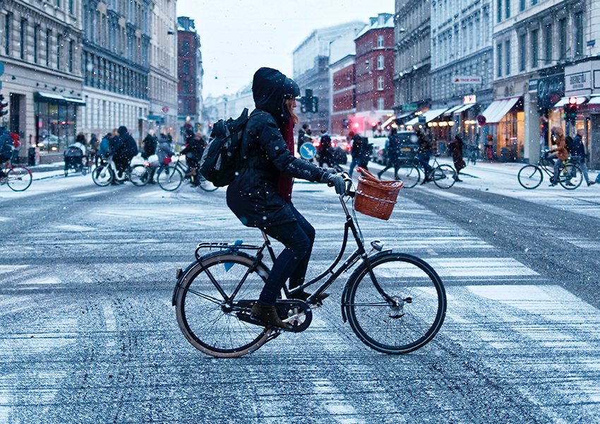 Eine Frau ist warm angezogen und fährt mit ihrem winterfesten Fahrrad über eine Straße, auf der etwas Schnee liegt.