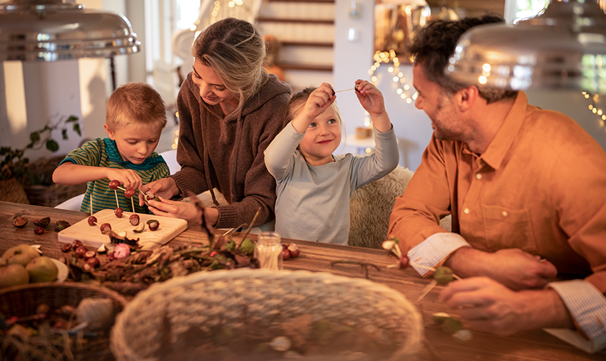 Mutter und Vater basteln mit ihren Kindern am Esstisch Kastanienmännchen.