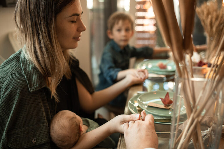Mutter mit Baby im Arm sitzt mit ihrer Familie am Tisch, sie hält die Händer der anderen und ist dankbar für das Essen.