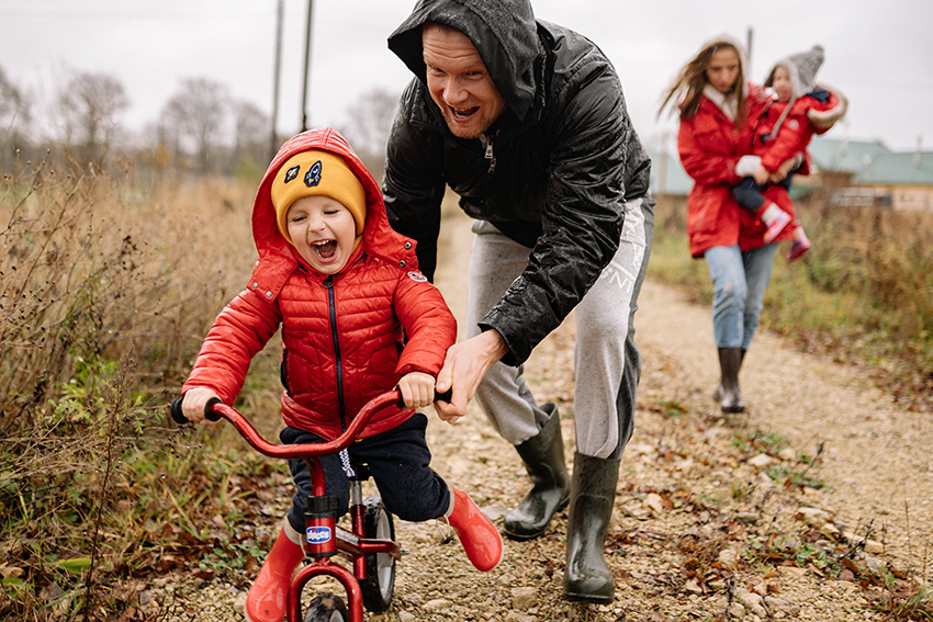 Ein Vater schiebt seinen kleinen Sohn auf dem Fahrrad an, die Familie trägt Regenklamotten.