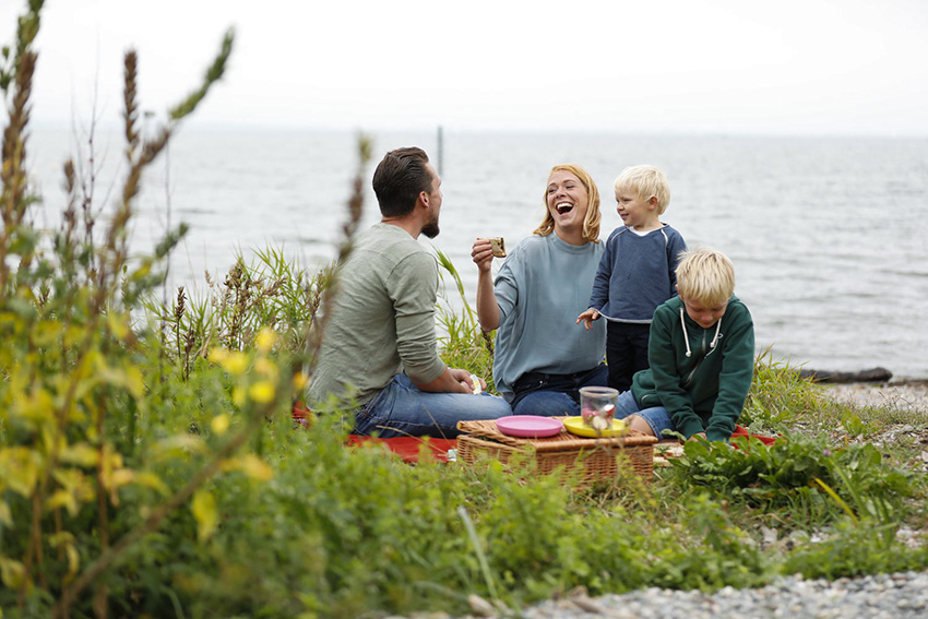 Eine Familie sitzt bei ihrem Mikroabenteuer auf einer Decke an einem Fluss und macht ein Picknick.