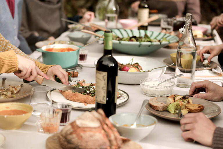 Ein langer Tisch ist gedeckt und mit Essen und Getränken beladen, um den Tisch sitzen Personen und essen gemeinsam.