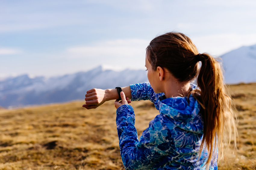 Mädchen steht auf einer Wiese mit Bergblick und misst ihren Puls auf einem Fitness-Armband.