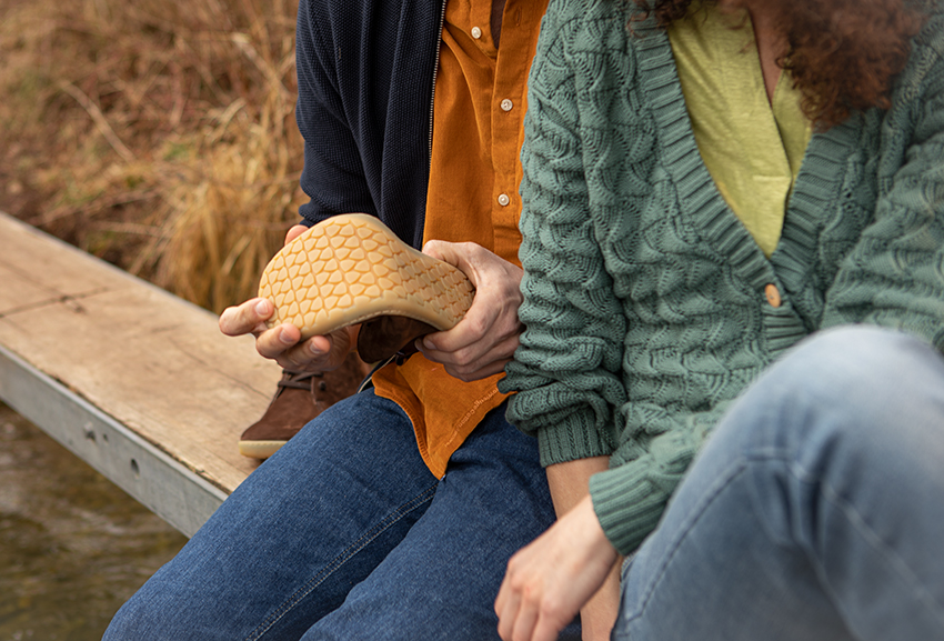 Ein Paar sitzt auf einem Holzsteg und biegt Barfußschuhe in den Händen.