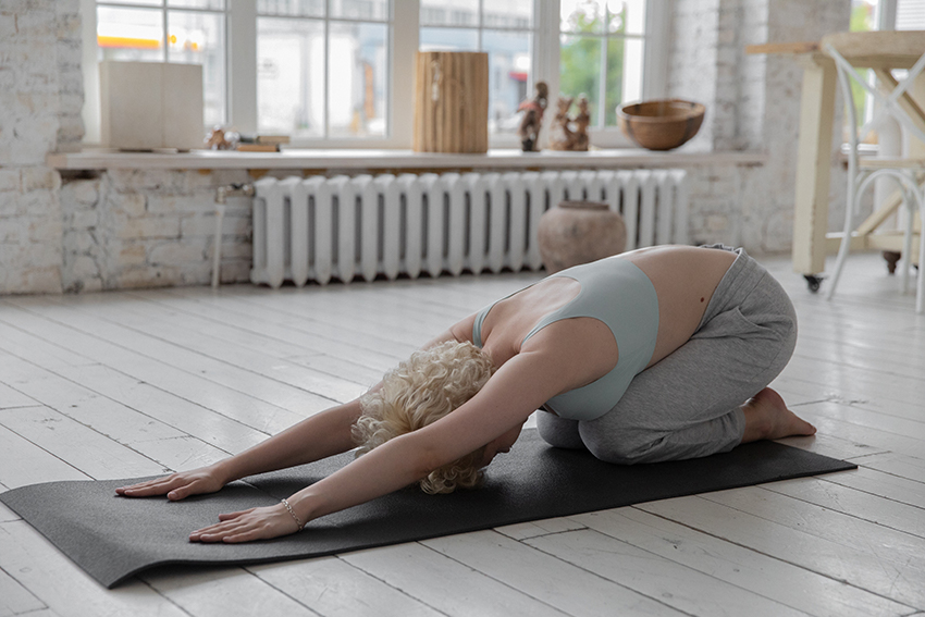 Eine Frau kniet in Sportklamotten auf einer Yogamatte, ihr Überkörper ist auf der Matte abgelegt, mit den Armen stützt sie sich auf der Matte ab.