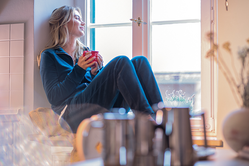 Eine Frau sitzt auf ihrer Fensterbank und schaut hinaus, dabei hält sie eine Tasse in den Händen.