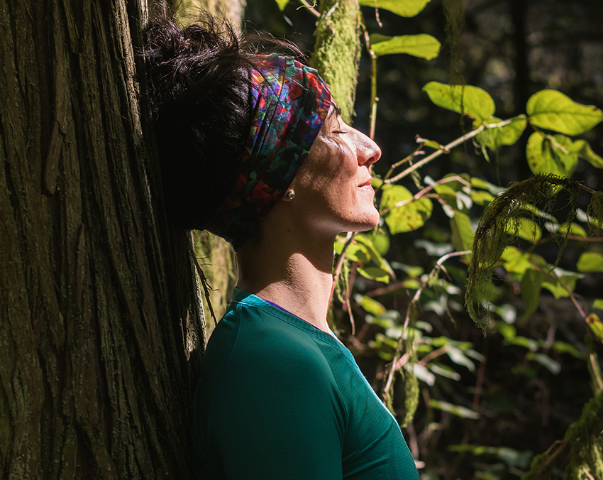 Eine Frau lehnt im Wald mit geschlossenen Augen an einem Baum und meditiert.