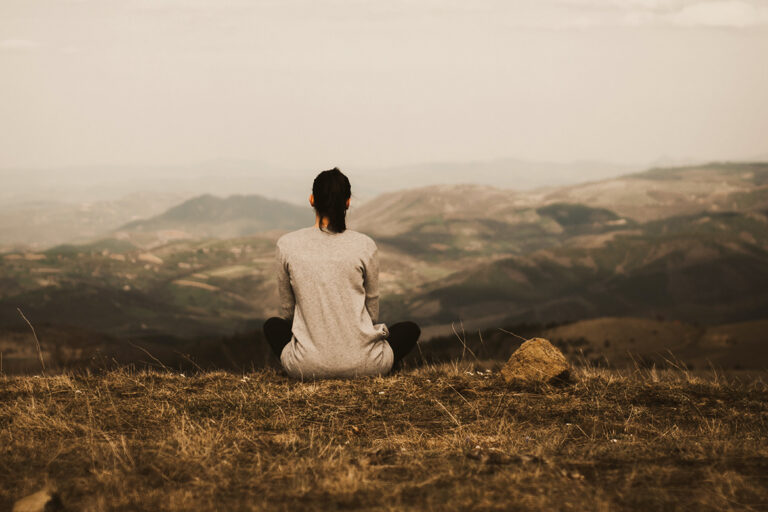 EIne Frau sitzt zur Meditation auf einer Wiese auf einem Berg und schaut entspannt ins Tal.