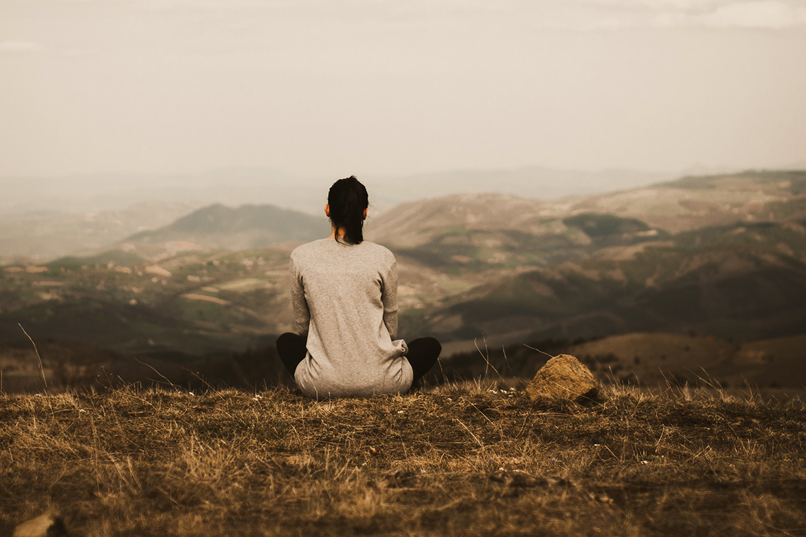 EIne Frau sitzt zur Meditation auf einer Wiese auf einem Berg und schaut entspannt ins Tal.