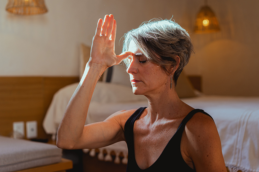 Frau sitzt mit geschlossenen Augen in ihrem Schlafzimmer, die Hand ist mit dem Daumen an die Stirn gelegt.