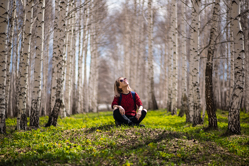 Eine Frau sitzt im Schneidersitz auf einer Wiese zwischen Birkenbäumen und hält ihr Gesicht der Sonne entgegen.
