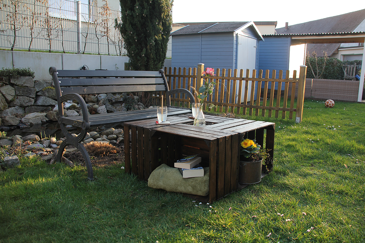 Der Gartentisch aus Holzkisten steht auf der Wiese vor einer Gartenbank.