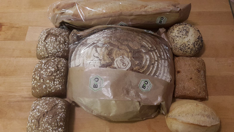 Ein großes rundes Brot liegt umgeben von Baguette und sechs Brötchen auf dem Holzbrett.