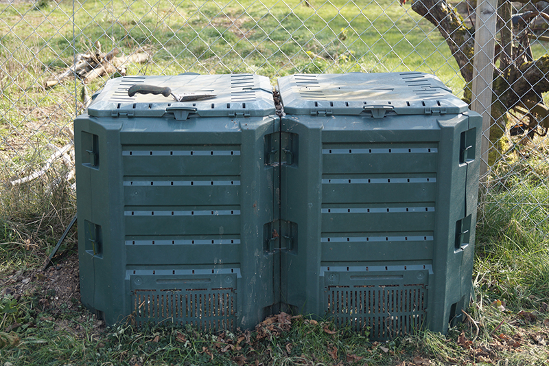 Twee donkergroene compostbakken staan naast elkaar bij een hek.