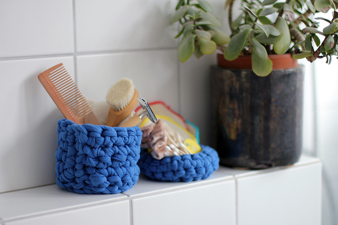 Blaues hohes und flaches Häkel-Utensilo aus Textilgarn im Badezimmer, befüllt mit plastikfreien Badutensilien.