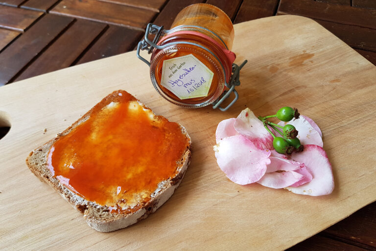 Auf dem Holzbrett liegt ein mit Butter und hellroter Hagebutten-Marmelade bstrichenes Brot. Im Hintergrund ein Glas mit dem Aufstrich und Rosenblüten.