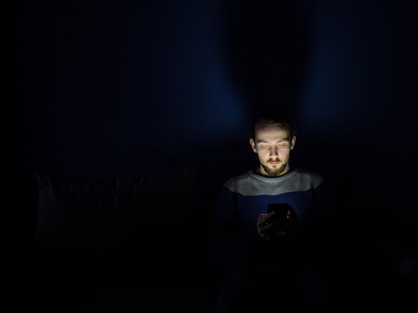 Ein Mann sitzt im Dunkeln in seinem Bett und wird von seinem Handy beleuchtet.