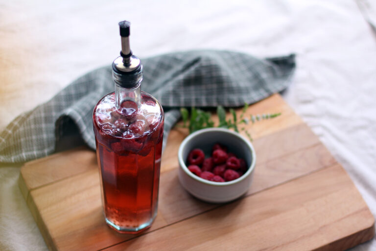 Eine Flasche selbst gemachter Himbeeressig steht auf einem Holzbrett, daneben steht eine Schale mit Himbeeren.