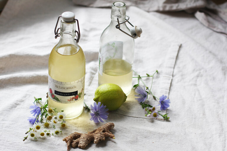 Zwei Flaschen Ingwerbier stehen nebeneinander, eine Ingwerknollen, Blumen und eine Limette liegen dabei.