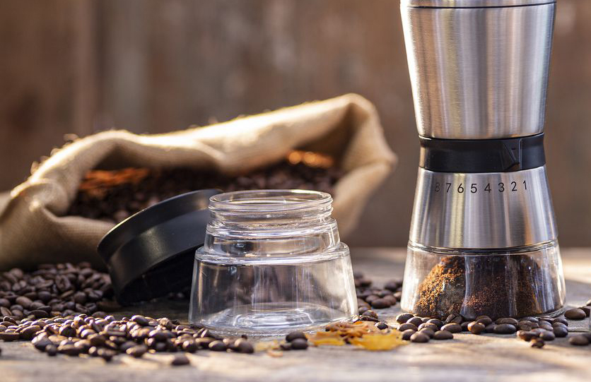 Kaffeebohnen liegen verstreut um eine kleine Kaffeemühle aus Edelstahl und Glas herum.