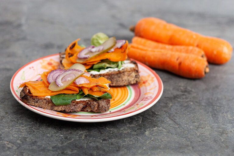 Neben dem Teller, auf dem Bauernbrot mit Karottenlachs , Salat und Zwiebelringen drapiert ist, liegen Karotten.