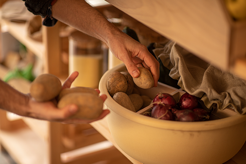 Kartoffeln und Zwiebeln werden in einem abgedeckten Topf gelagert, aus welchem Kartoffeln durch zwei Hände entnommen werden. 