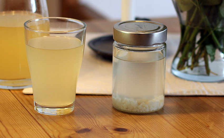 Das Glas mit den weißen Wasserkefir-Kristallen steht auf dem Tisch neben einem Glas mit fertigem Kefir.