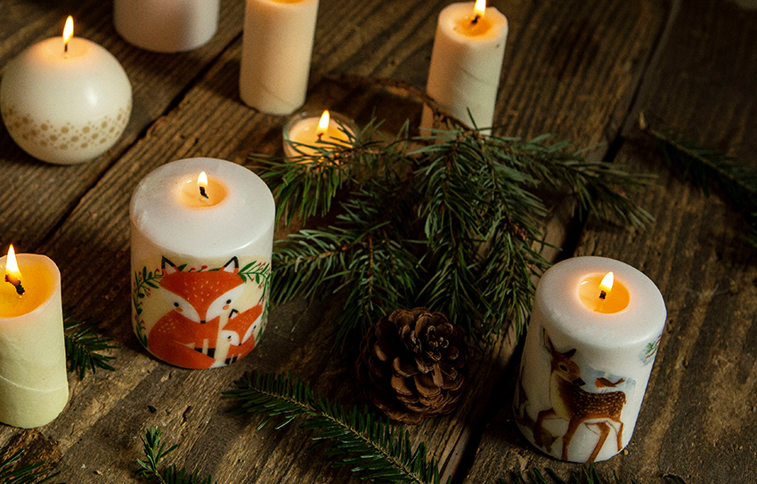 Selbst gemachte und verzierte Kerzen stehen leuchtend auf einem Holztisch mit Tannenzweigen.