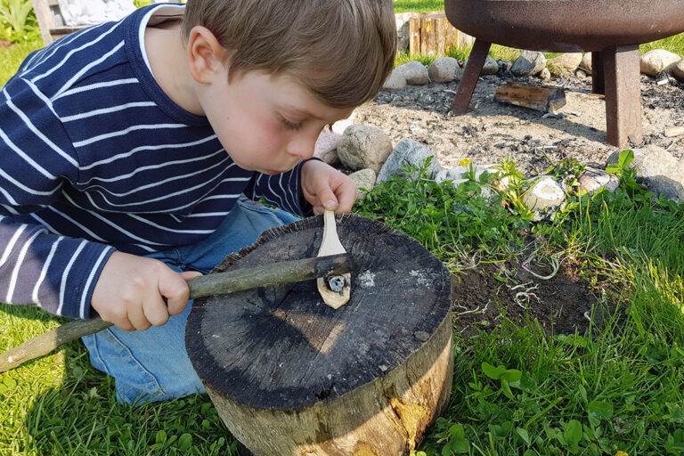 Ein Junge sitzt vor einem Holzblock und brennt mit Stock und Glut eine Mulde in einen geschnitzten Löffel.