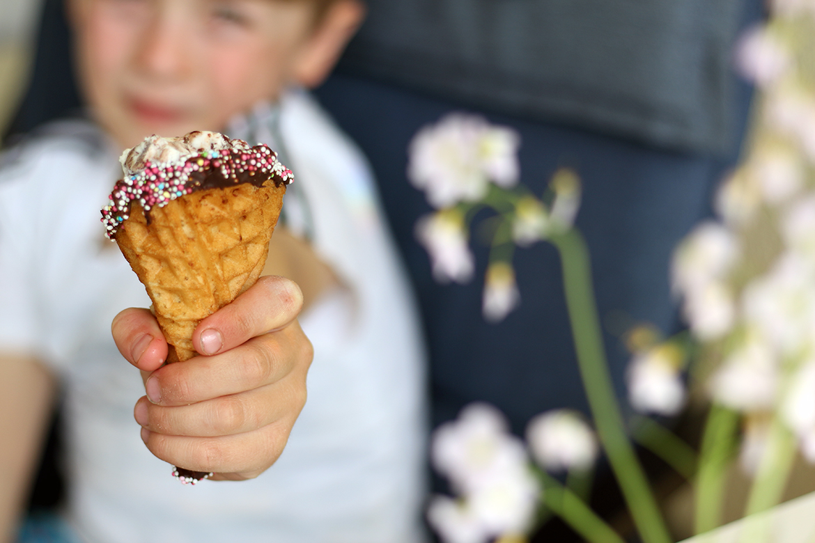 Ein Kind hält eine selbst gemachte Eiswaffel in die Kamera, die Waffel ist mit am Rand mit Schokolade und Streuseln verziert.